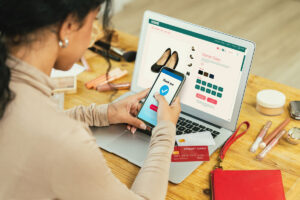 Woman making an online payment via smartphone while shopping for women's shoes on a laptop, surrounded by cosmetics and credit cards on a wooden desk